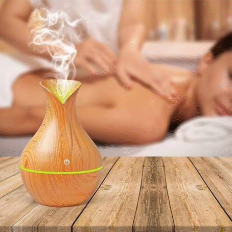 The image shows a wood-grain aroma diffuser emitting gentle steam, placed on a wooden table. In the blurred background, a woman is receiving a relaxing massage at a spa, highlighting the product's use for creating a calming and relaxing atmosphere.