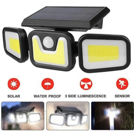 In the photo, an outdoor LED lamp with a solar panel is mounted on the wall of a building. The lamp features three adjustable light panels emitting a bright white light, illuminating the surrounding area at night. In the background, the blurred outlines of other buildings can be seen in the dark.