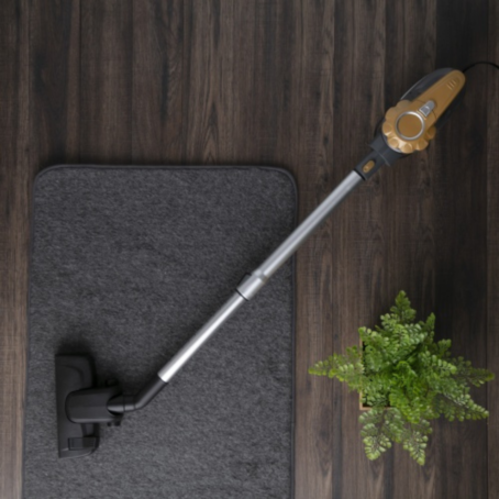 top view of a grey and gold stick vacuum cleaner cleaning a dark carpet on wooden floor, next to a green potted plant