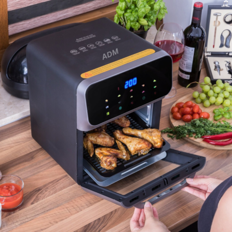 The image shows a modern ADM oven on a wooden kitchen countertop. Golden chicken wings are being baked inside the oven. In the background, there are fresh vegetables, wine, and herbs, emphasizing a homey atmosphere and healthy cooking.