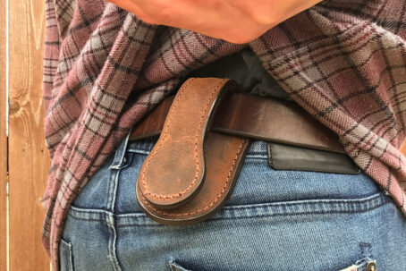 Close-up of a person in a plaid shirt and jeans wearing a brown leather coin pouch attached to their belt.